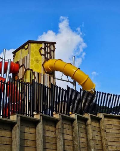 Low-angle view of a yellow tube slide and honeycomb-panelled timber play tower against a blue sky, set on a timber-retained bank